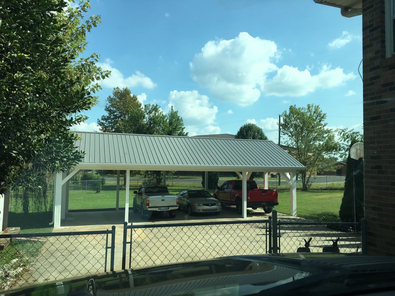 A covered parking area with three vehicles: a white truck, a gray sedan, and a red truck. The structure has a metal roof and is situated in a fenced yard with trees and a clear, partly cloudy sky in the background. from Higley Construction