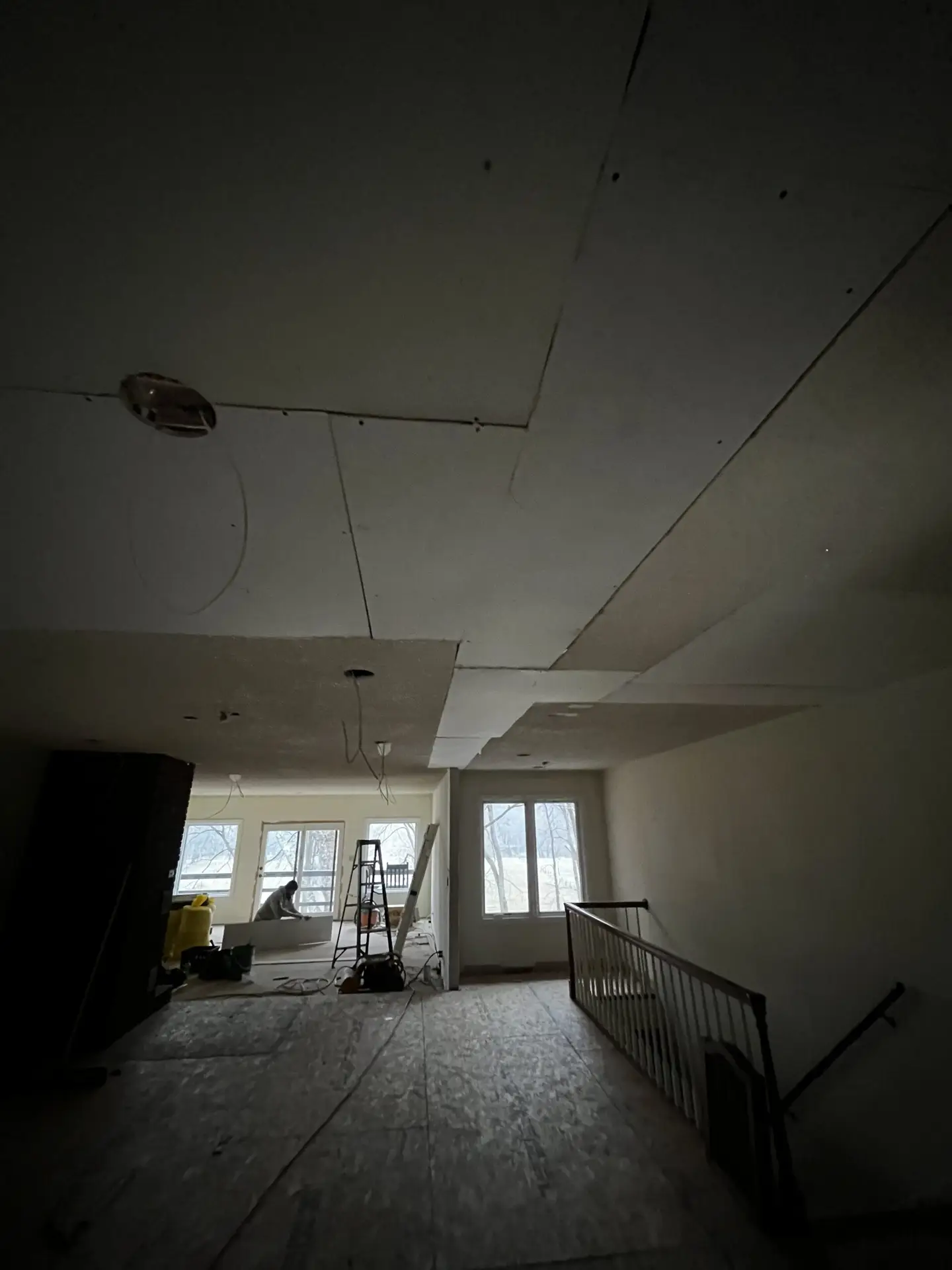 A dimly lit room under renovation, with missing ceiling panels and exposed wiring. A ladder stands beneath an incomplete ceiling, and a window at the far end lets in faint natural light. Plastic sheeting covers the floor next to a staircase with a black railing. from Higley Construction