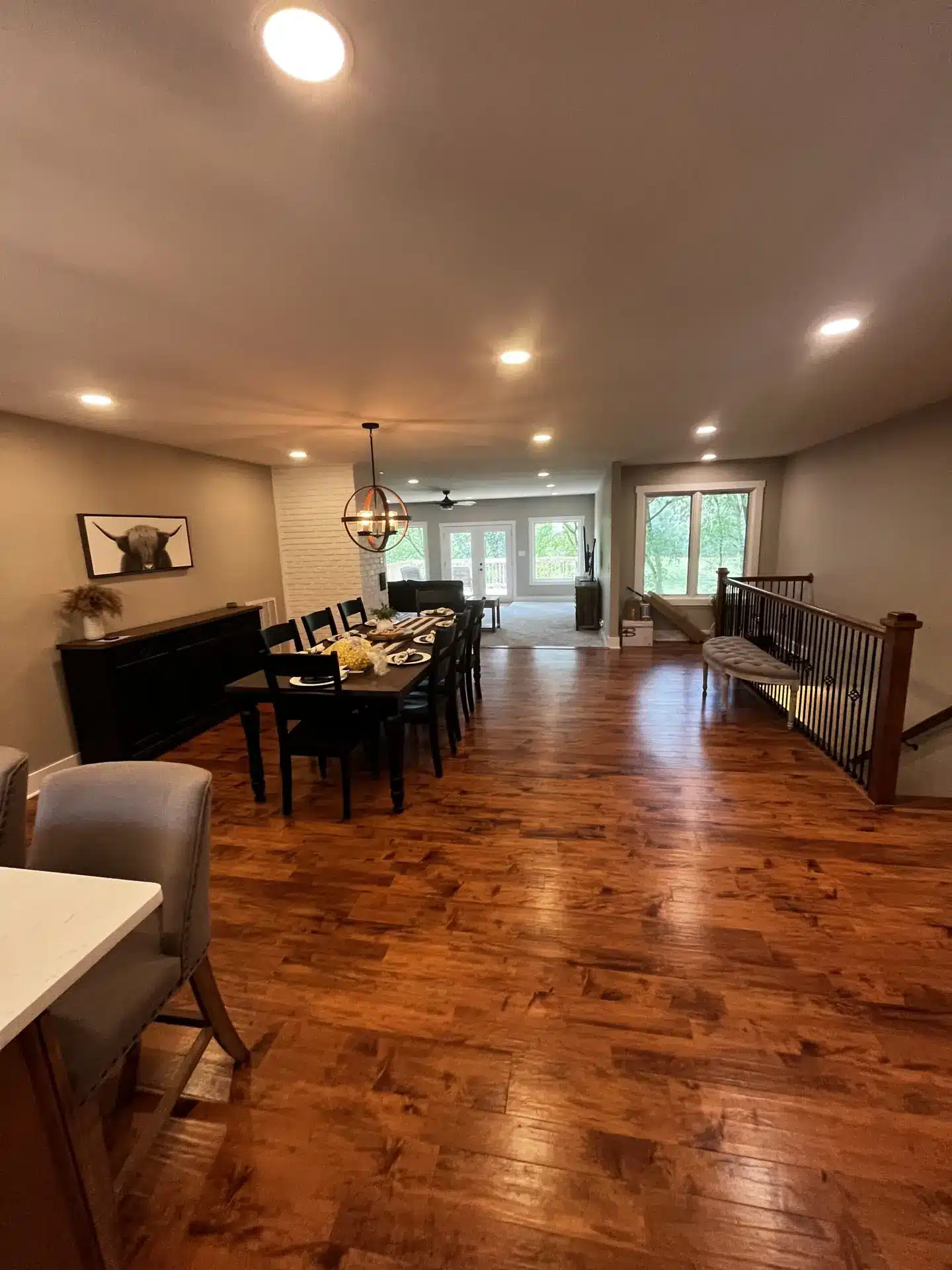 A spacious dining area with hardwood floors features a long table set for a meal, surrounded by chairs. There's a kitchenette with grey barstools on the left and a large window at the back letting in natural light. A round light fixture hangs over the table. from Higley Construction