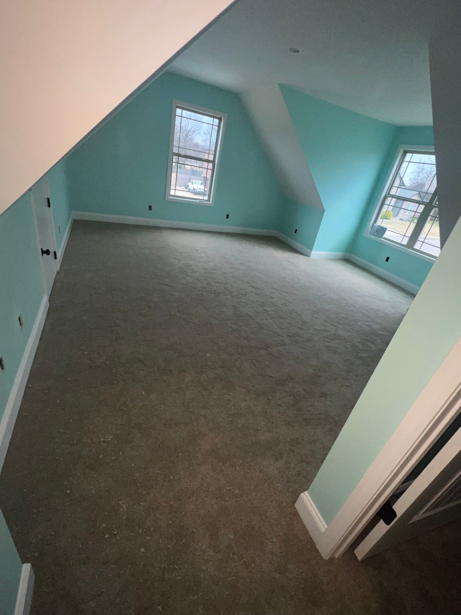 An empty room with light blue walls, two large windows, and light grey carpeting. The ceiling slants down on one side, and the room has white baseboards and trim. Two walls have electrical outlets and switches, and one wall has a closed door. from Higley Construction