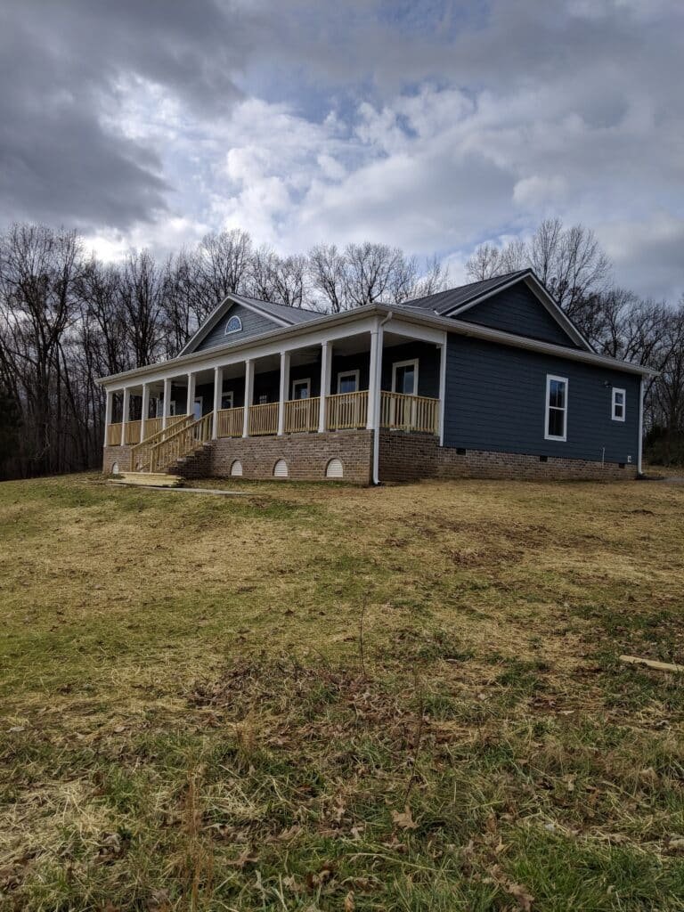 A single-story house with a blue exterior and a long wooden porch set against a backdrop of leafless trees and a cloudy sky. The house sits on a large grassy lawn that gently slopes upwards. from Higley Construction