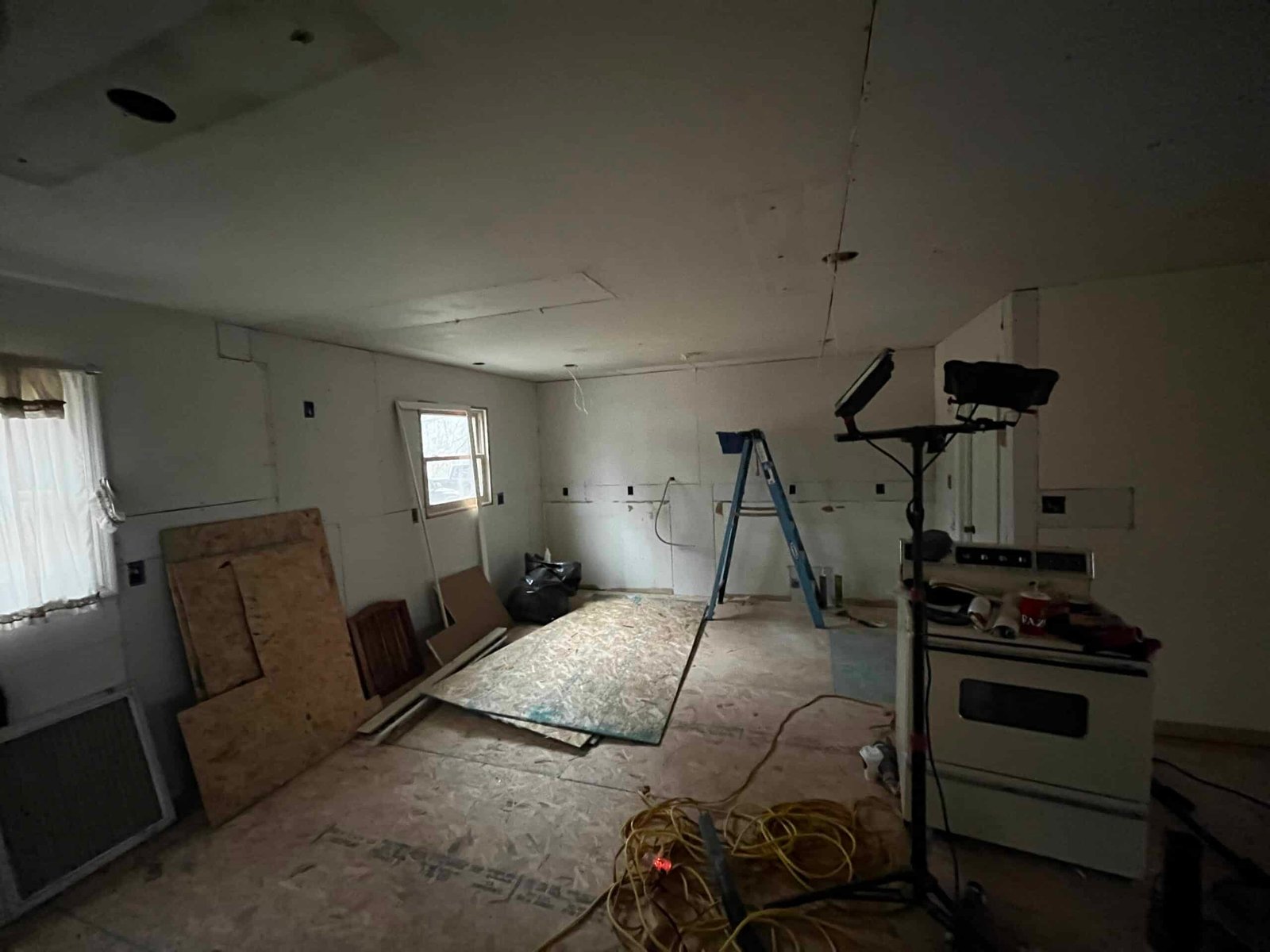 A dimly lit, partially renovated room with construction materials scattered around. An unassembled ceiling, an upright ladder, an uninstalled oven, a stack of plywood, and coiled extension cords are visible. Two windows provide minimal natural light. from Higley Construction