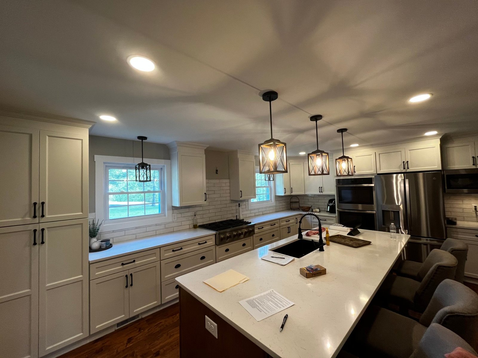 A modern kitchen with white cabinets and a large central island. Three pendant lights hang above the island, which has seating and papers on top. Stainless steel appliances are visible in the background, and a small potted plant sits near a window on the left. from Higley Construction