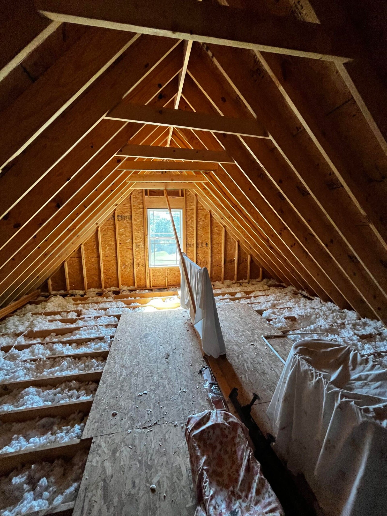 An unfinished attic space with visible wooden beams, insulation material on the floor, and a single window at the far end. Some items are covered with white cloths, and long sheets hang from the ceiling. Sunlight filters through the window, illuminating the area. from Higley Construction