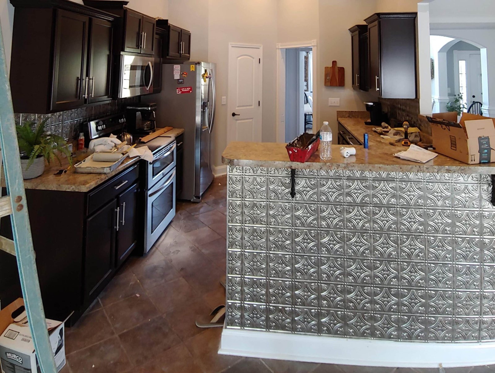 A modern kitchen with dark cabinets, stainless steel appliances, and a decorative backsplash. The countertop has various items, including a water bottle, tools, and a small red toolbox. A ladder is seen on the left, and there are cardboard boxes on the right. from Higley Construction