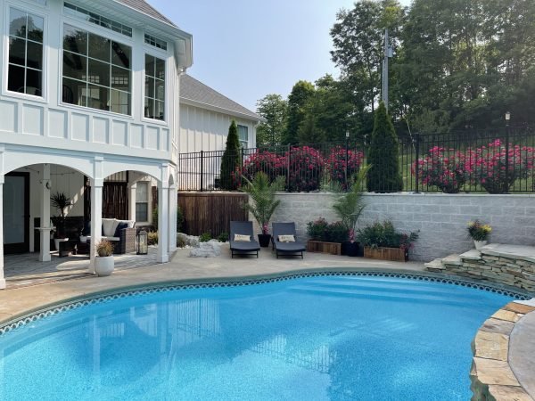 A backyard with a bright blue swimming pool, bordered by a stone patio. Two lounge chairs with cushions are placed poolside. The house has large windows and an upper balcony. A garden with flowering bushes and trees surrounds the pool area. from Higley Construction