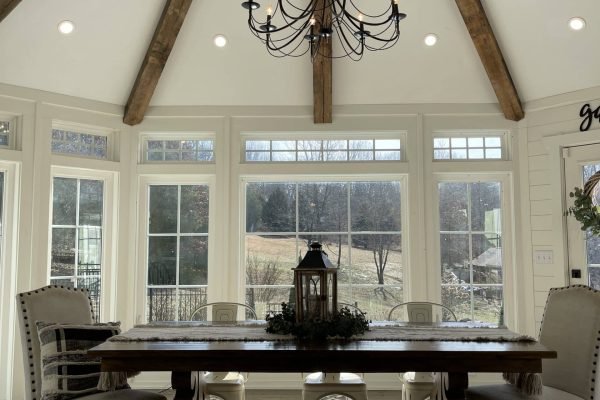 A dining room features a large wooden table with two benches and end chairs. A centerpiece with greenery and a lantern sits on the table. The room has a high, vaulted ceiling with exposed wooden beams and a black chandelier. Sunlight streams through large windows. from Higley Construction