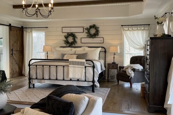 A cozy bedroom with a rustic theme features a metal-framed bed adorned with white and gray bedding. Two wreaths hang above the bed. The room includes wooden beams on a white ceiling, a chandelier, a sliding barn door, an armchair, and a dresser. Light streams through sheer curtains. from Higley Construction