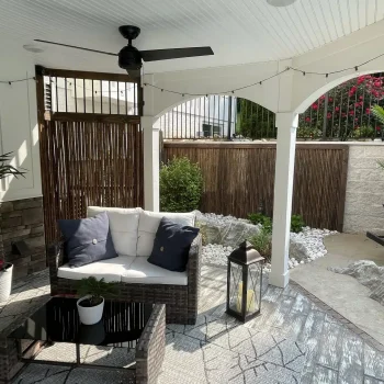 A cozy outdoor patio features a wicker sofa with white cushions and dark accent pillows, a glass-top coffee table, and a reclining lounge chair. String lights hang under the roof, and there are potted plants, bamboo privacy screens, and a lantern in the space. from Higley Construction