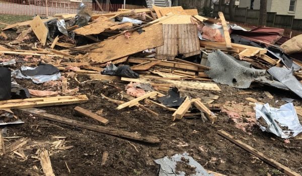 A pile of debris, including wood, metal, and insulation, is scattered on the ground in front of a partially demolished house. The house in the background appears intact, with a staircase leading to the second floor. Bare trees are visible around the area. from Higley Construction