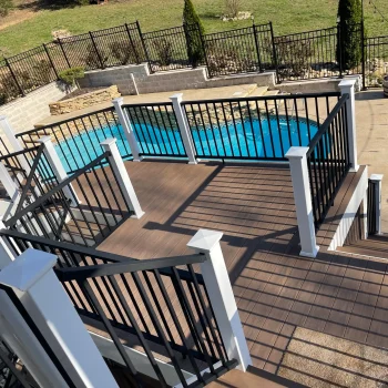 A multi-level wooden deck with black railings overlooks a swimming pool in a fenced backyard. The deck has several stairs leading down to the pool area, which is adjacent to a stone patio. Trees and grass are visible in the background. from Higley Construction