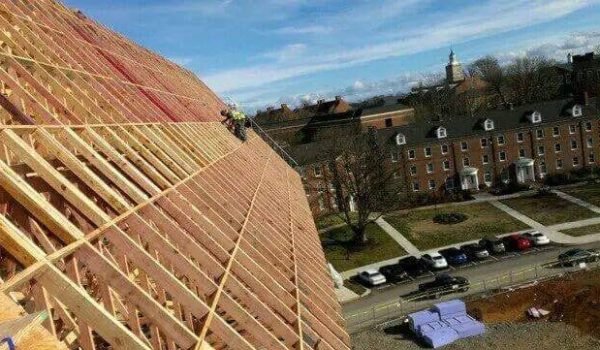 Workers in safety gear are on a steep wooden frame of a large roof under construction. Below, a row of cars is parked beside a brick building with dormer windows. The sky is clear, and the area has trees and lawns, suggesting a campus or institutional setting. from Higley Construction