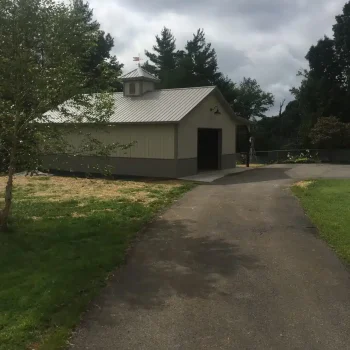 A path leads to a small, light-gray building with a metal roof and a cupola on top. The building is situated among green grass and trees under a cloudy sky. from Higley Construction