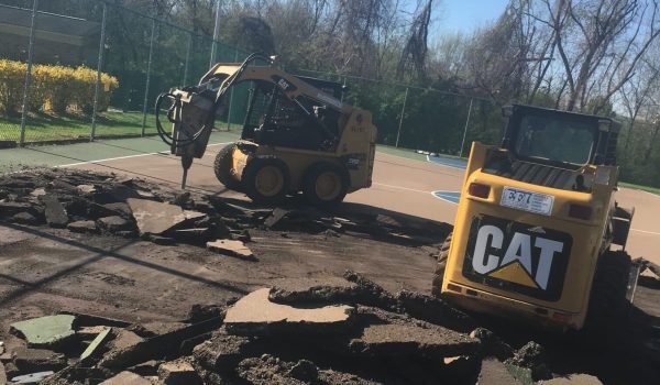 Workers using heavy machinery to break and remove the asphalt of an outdoor basketball court surrounded by a fence and trees, with clear blue skies in the background. The machinery has "CAT" branding visible. from Higley Construction