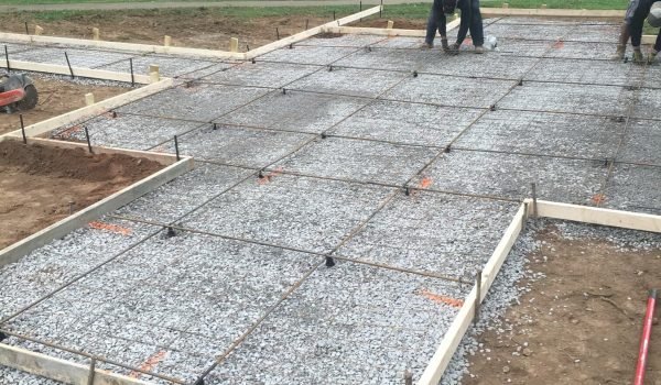 Construction site with wooden frames outlining a rectangular area filled with gravel. Metal rebar grids are laid on top of the gravel for reinforcement. Two workers are bending over the grids, adjusting the rebar, while tools and materials are scattered nearby. from Higley Construction