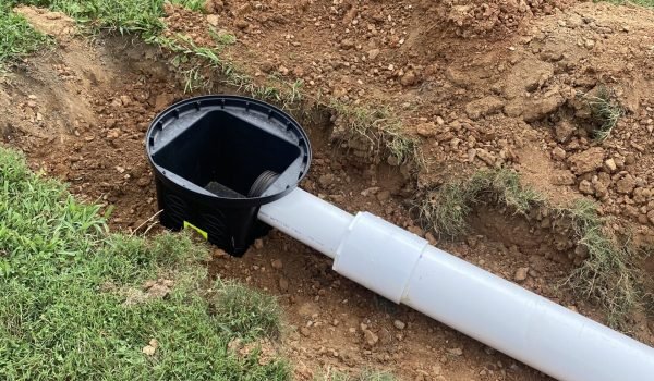 A partially buried drainage pipe with a black, hexagonal catch basin at the joint. The surrounding area has freshly turned soil and patches of grass, indicating recent installation or maintenance work. The scene is set outdoors on a grassy surface. from Higley Construction