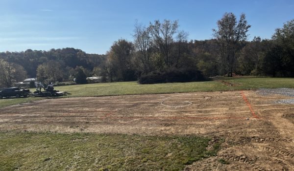 A construction site on a sunny day, with a cleared dirt foundation marked with orange spray paint. Equipment and vehicles are parked on the left, and there are trees and grassy areas in the background. from Higley Construction