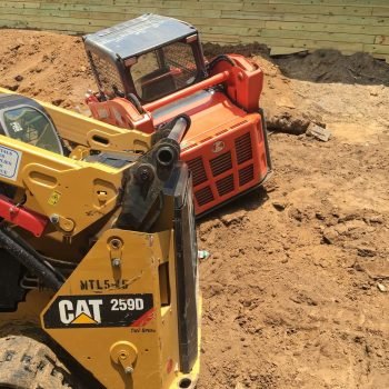 A construction site shows a yellow Caterpillar 259D skid steer positioned next to an overturned orange skid loader on a dirt surface. The background features a wooden retaining wall under construction with various construction materials scattered around. from Higley Construction