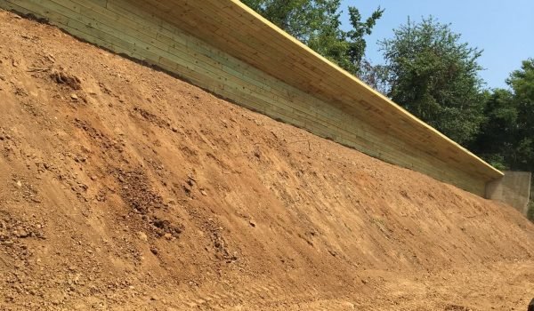 A large, recently excavated slope of dirt is supported by a retaining wall at the top. The wall is made of large wooden beams and is situated in front of a backdrop of green trees and a clear blue sky. Construction machinery tracks are visible on the dirt. from Higley Construction