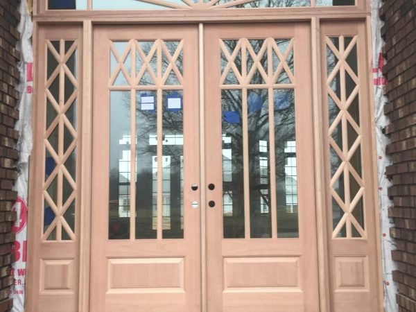 A wooden double door with intricate cross-hatch design details is set within a brick building. The door has glass panels on each side and an arched transom window above. Construction materials and 84 Lumber branding are visible around the doorway frame. from Higley Construction