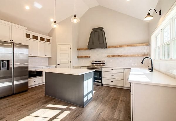 A spacious, modern kitchen with white cabinetry, a stainless steel refrigerator, and a gray central island. It has wooden floors, a white tile backsplash, and open wooden shelves. Three pendant lights hang from the ceiling, and the room is brightly lit by natural light. from Higley Construction