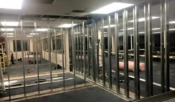 A partially constructed building interior with exposed metal framing for walls. Several ceiling tiles are missing, revealing wires and structure above. Construction materials and equipment are scattered on the floor. Fluorescent lights are on overhead. from Higley Construction