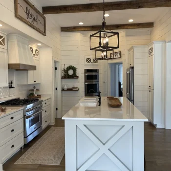 A modern farmhouse kitchen with white cabinetry, a large center island with a white countertop, stainless steel appliances, and wooden ceiling beams. A rustic light fixture hangs above the island, and a sign reading "blessed" is on the wall. from Higley Construction