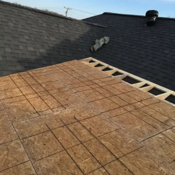 A rooftop under construction with partially installed plywood sheathing. The adjacent section has completed shingles, and some construction materials are visible. A clear blue sky is in the background. from Higley Construction